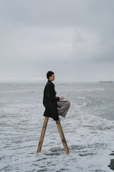 A person on a ladder by the sea, working on a laptop, symbolizing balance between technology and nature.